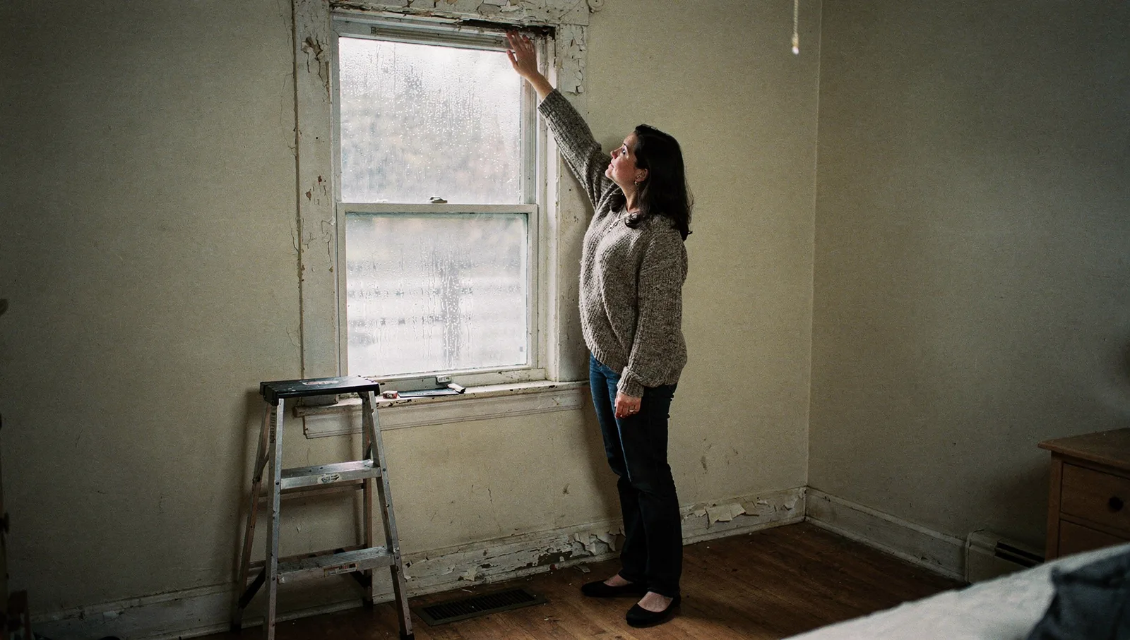 Woman inspecting moisture damage and peeling paint around a window frame inside an older apartment.